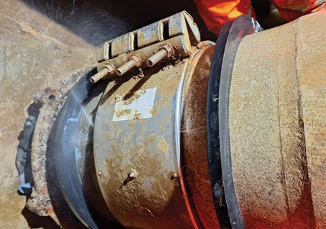 Grande conduite métallique présentant une fuite et une pince de réparation rouillée sur laquelle de l'eau s'écoule visiblement.